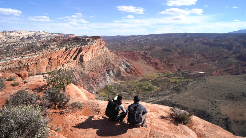 Two people sit on a cliff overlooking a wide desert canyon with layered red rock in Utah