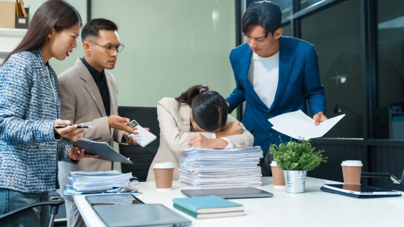 A stressed team surrounds a leader with head down at a desk, showing communication breakdown linked to founder burnout