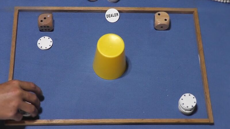 A Chō-Han dice game is set up with two dice, betting chips, and a covered cup on a blue mat