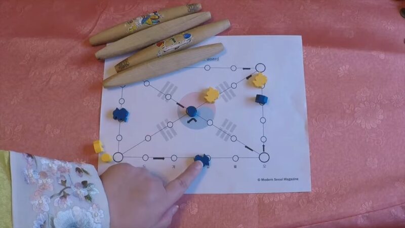 A person moves a game piece on a Yut Nori board with wooden throwing sticks beside it