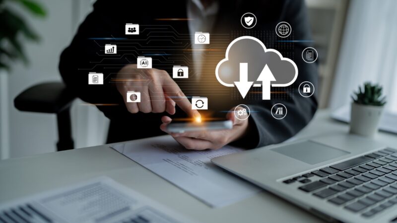 Person uses a smartphone beside a laptop with cloud transfer and security icons.
