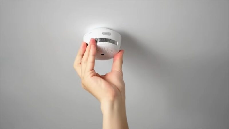 A hand checks a smoke detector on the ceiling.